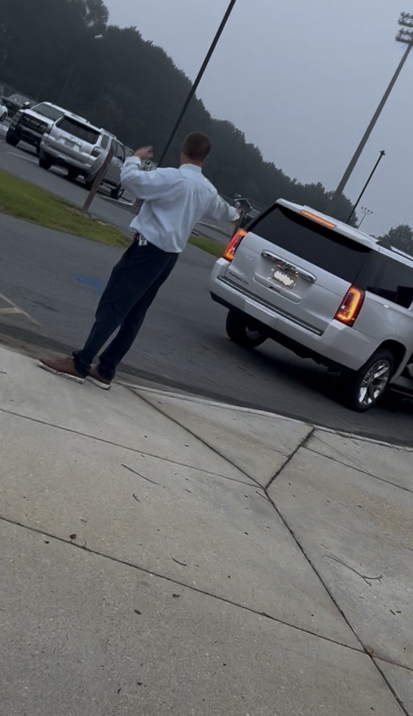 School principal is staying standing with navy pants and a white button up as he directs traffic as a white SUV passes by