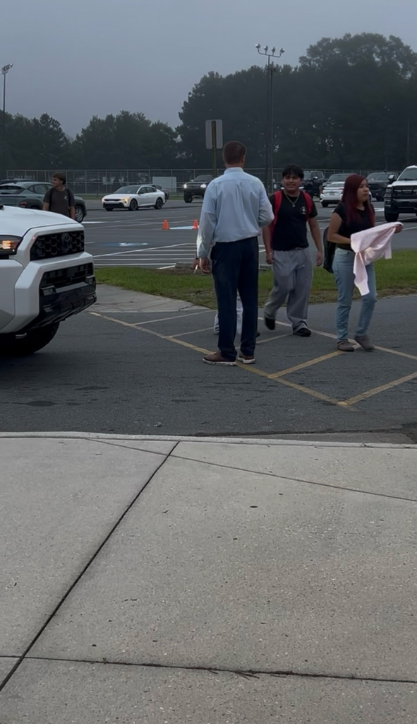 School principal in navy pants and a white button up can be seen blocking traffic as students cross in the crosswalk