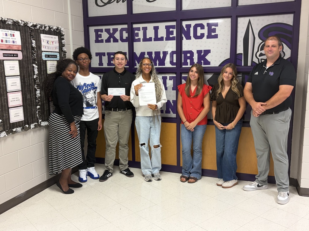 School staff, students, and representatives pose together in front of an Excellence and Teamwork wall graphic.