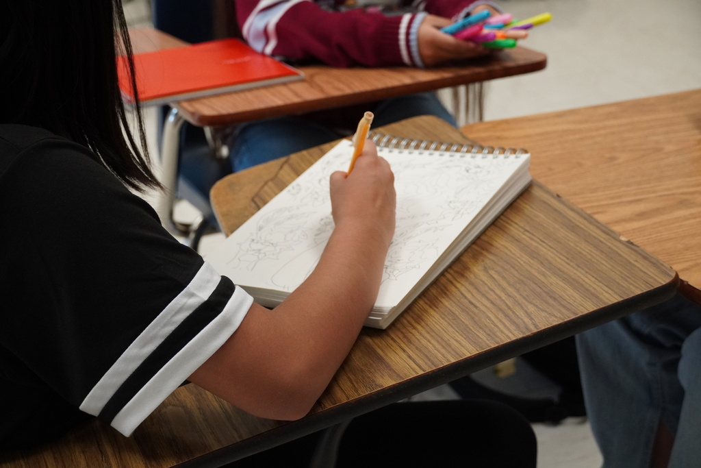 A student is drawing in a sketch pad while sitting at a desk. 