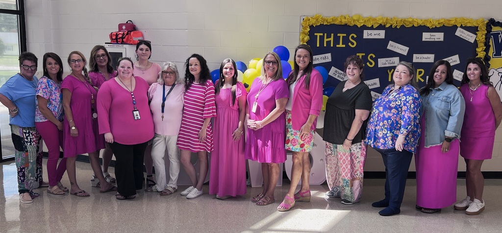 Alt text: A group of Bladenboro Middle School staff members pose together in the school atrium wearing shades of pink in remembrance of Dolly Parton. Blue and gold balloons and a school bulletin board are visible in the background.