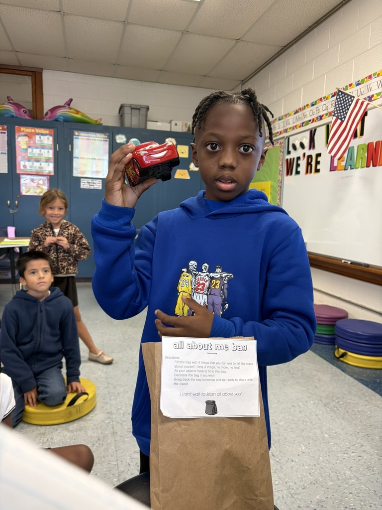 Student in a blue sweatshirt shares a toy car from his “All About Me” bag with classmates.