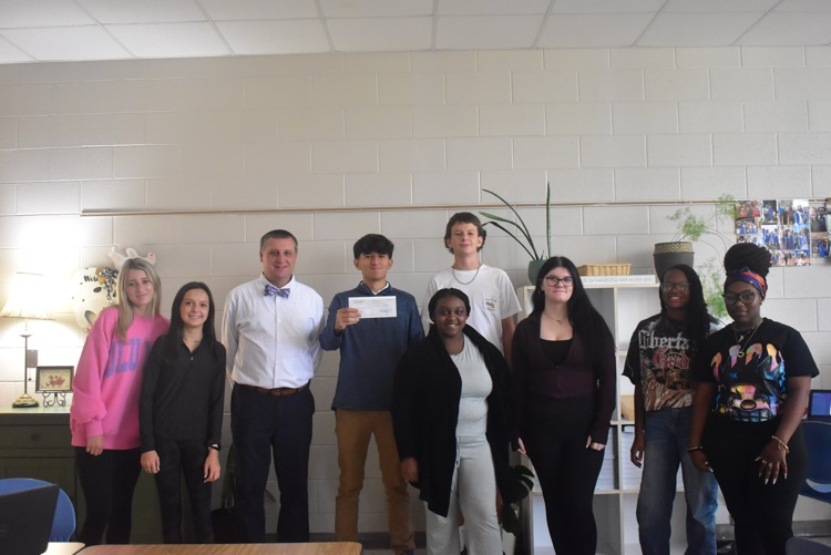 students and adult stand in front of a white wall. There is a lamp in the background a bookcase with plants and one student stands in the middle, holding a check.