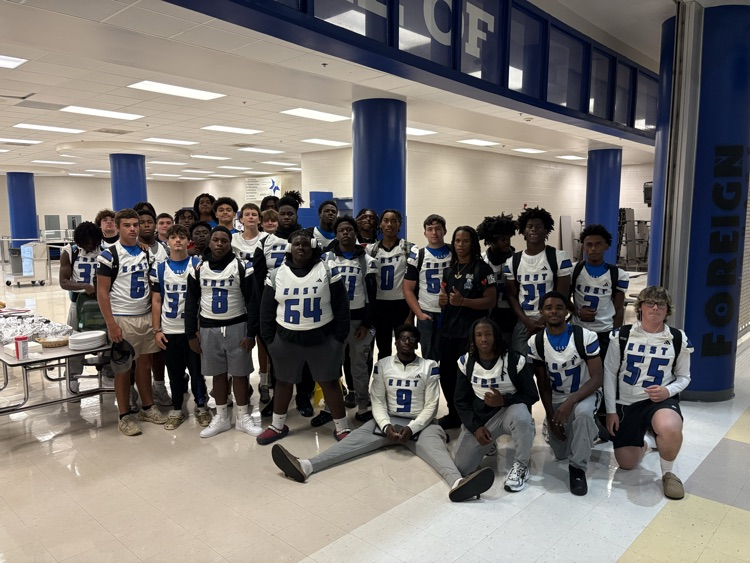 A large group of football players standing in a cafeteria with white jerseys that say East and their numbers on it there’s a table to the left that has food on it