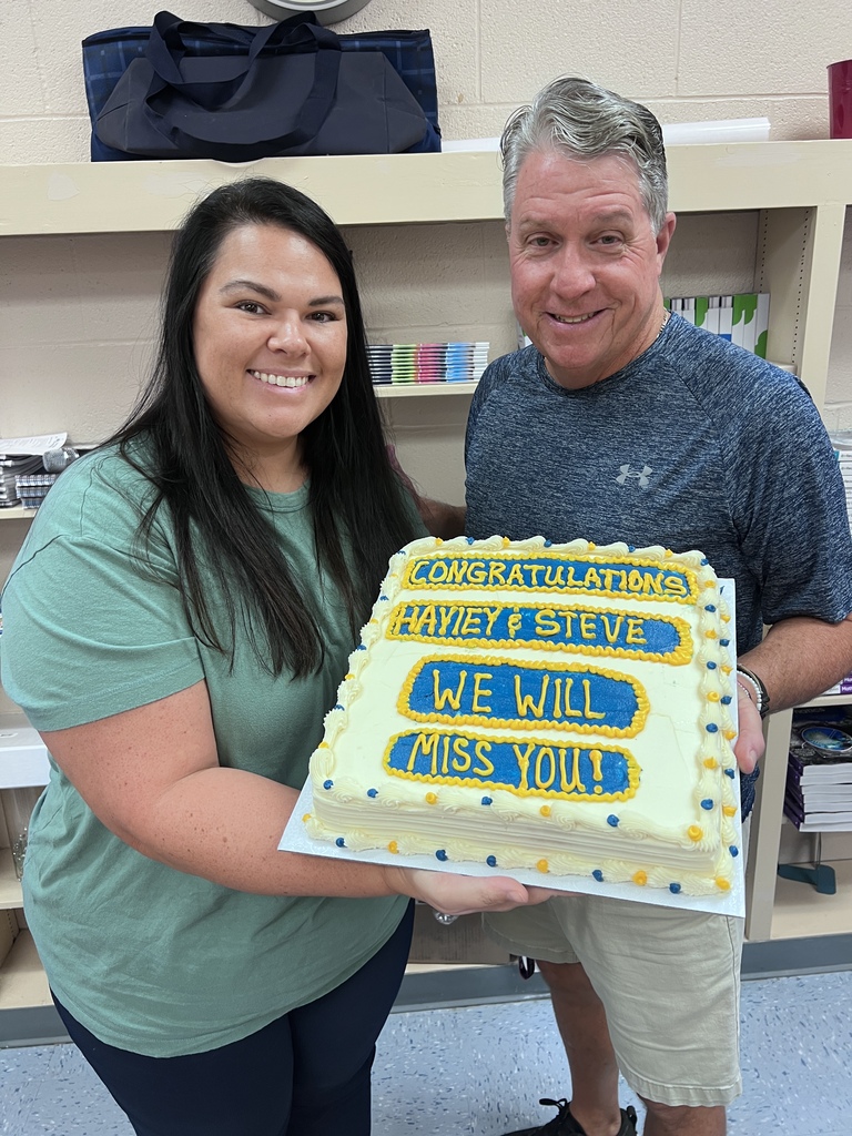Two Bladenboro Middle School staff members smile while holding a large blue, gold, and white cake that reads, “Congratulations Hayley & Steve. We will miss you!” during a celebration honoring their next chapters.