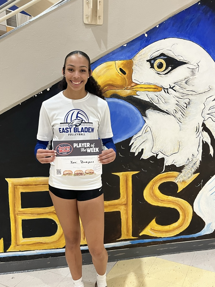 student standing in front of a wall with an eagle on it in black shorts, white T-shirt, holding a certificate