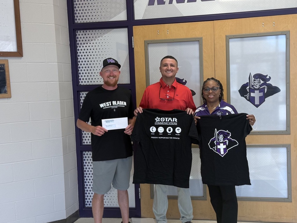 Three adults stand indoors holding black t-shirts in front of the school doors. Coach Jackson holds a check while the STAR rep and Mrs. Elliott hold the custom tshirts