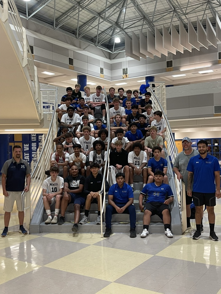 A group of 38 soccer players sit on a stairwell as three coaches stand beside them in the atrium of a school. One team is wearing white jerseys. The other team is wearing royal blue jerseys.