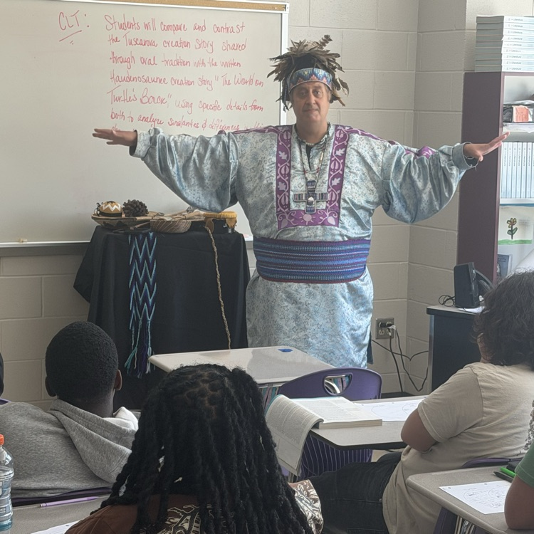 Indigenous speaker in traditional regalia stands before a classroom with arms outstretched during a presentation to students