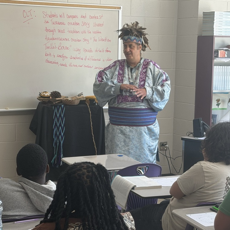 Indigenous speaker wearing traditional regalia and a feather headdress gestures with hands while speaking to classroom students