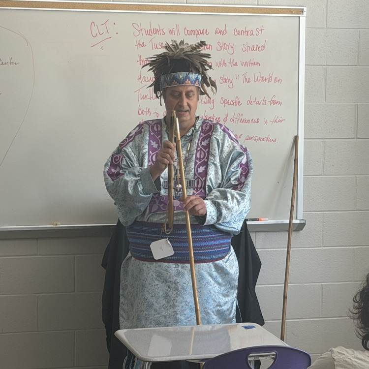 Indigenous presenter in traditional regalia, feather headdress, and patterned tunic holds bamboo flutes in front of a classroom whiteboard.