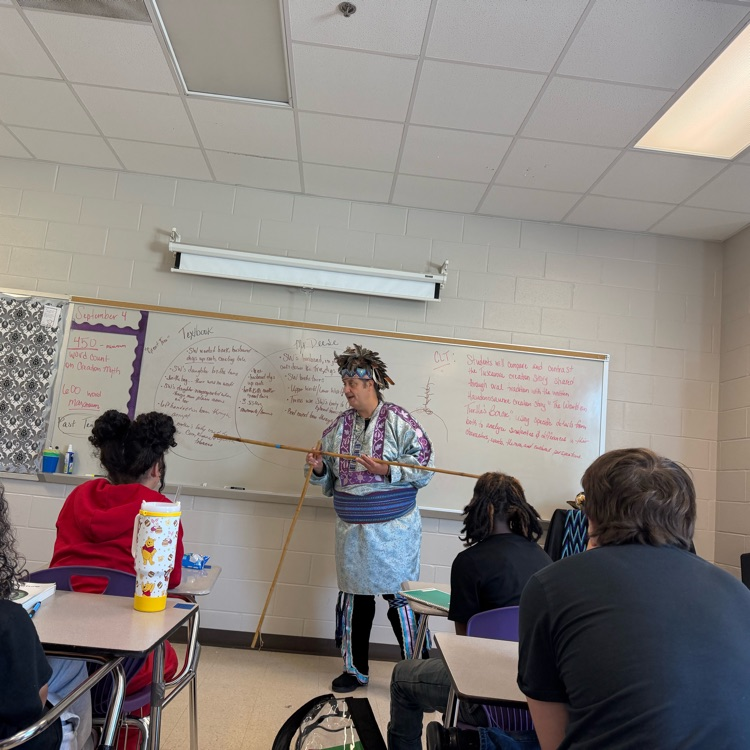 Wide view of a classroom with seated students watching an Indigenous speaker in traditional regalia hold a long wooden stick.