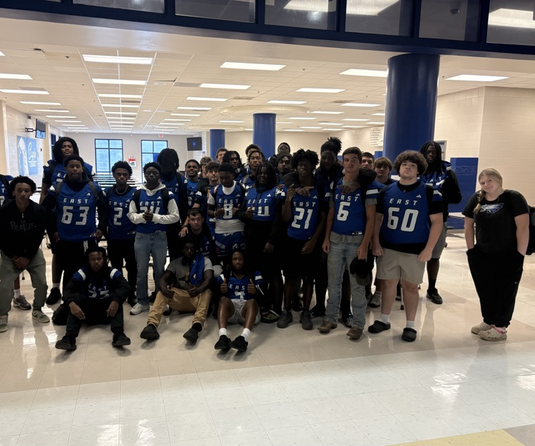 football player sitting and standing in a circle as they pose for a photo before they eat a pregame meal. Players are wearing East Bladen jerseys
