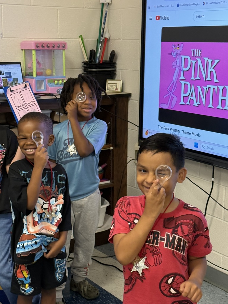 Three smiling students pose with magnifying glasses and detective badges during a library activity.