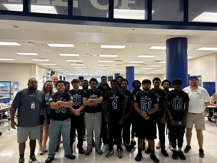 Group of football players stand with their arms crossed as a pose for a group photo with their coaches and the three people that provided them a pregame meal. They are standing at the entrance of the cafeteria.
