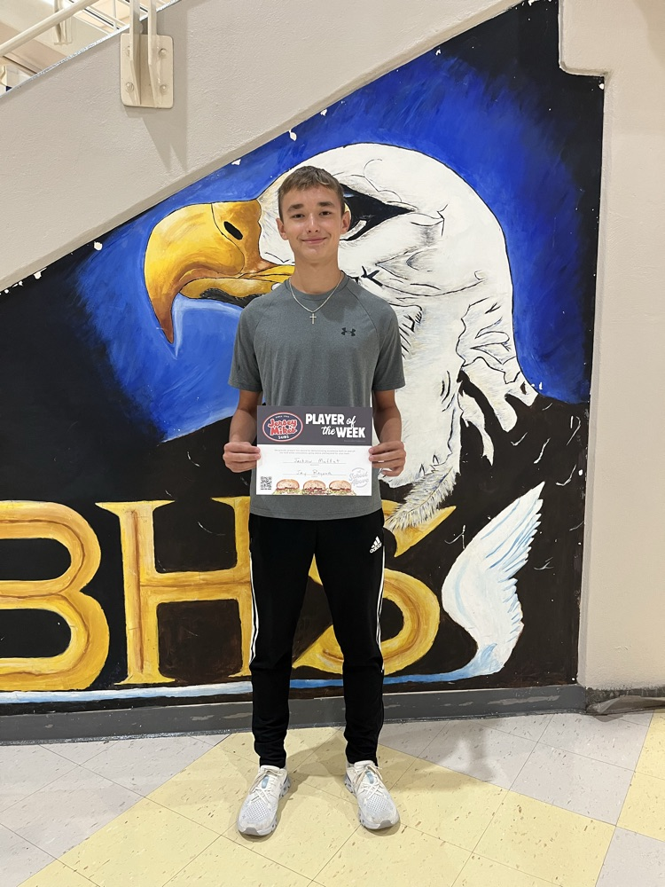 student and black pants, gray T-shirt, holding a jersey Mike’s certificate in front of an EBHS eagle backdrop