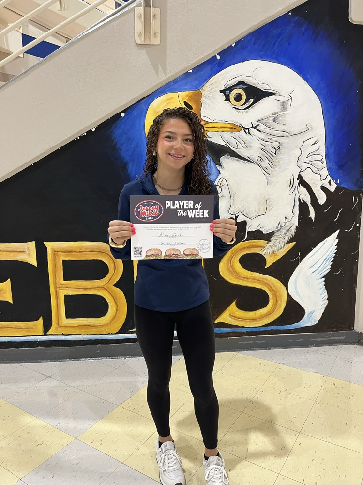 student and black pants, gray T-shirt, holding a jersey Mike’s certificate in front of an EBHS eagle backdrop