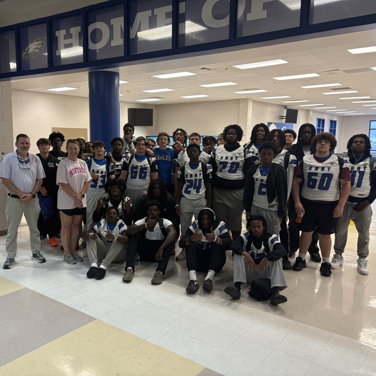 Group of football players standing with a man in khaki pants and a lady in a pink shirt in front of the cafeteria