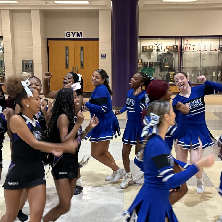 A group of cheerleaders in blue and black uniforms dance and jump excitedly in a tiled multi-story school lobby, with signs in the background for a gym and cafeteria.