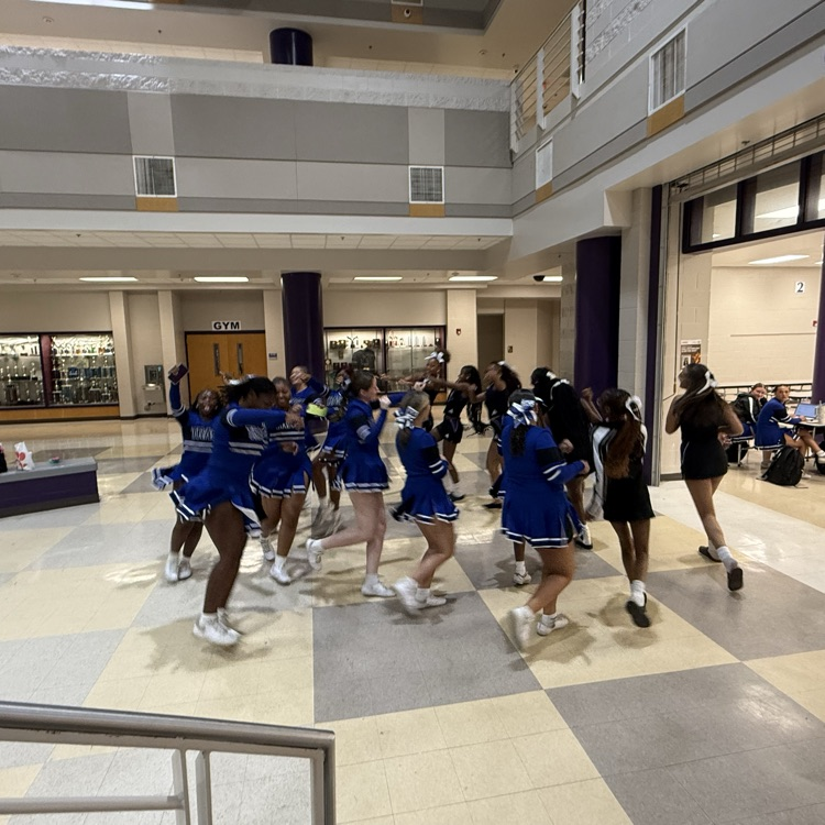 A group of cheerleaders in blue and black uniforms dance and jump excitedly in a tiled multi-story school lobby, with signs in the background for a gym and cafeteria.