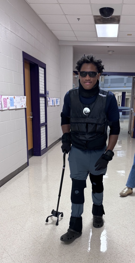 A student wears a weighted geriatric simulation suit, dark glasses, joint restraints, and holds a quad cane