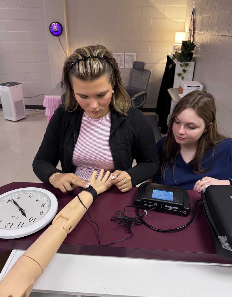 Two students practice medical training by examining a training arm connected to a Realityworks device