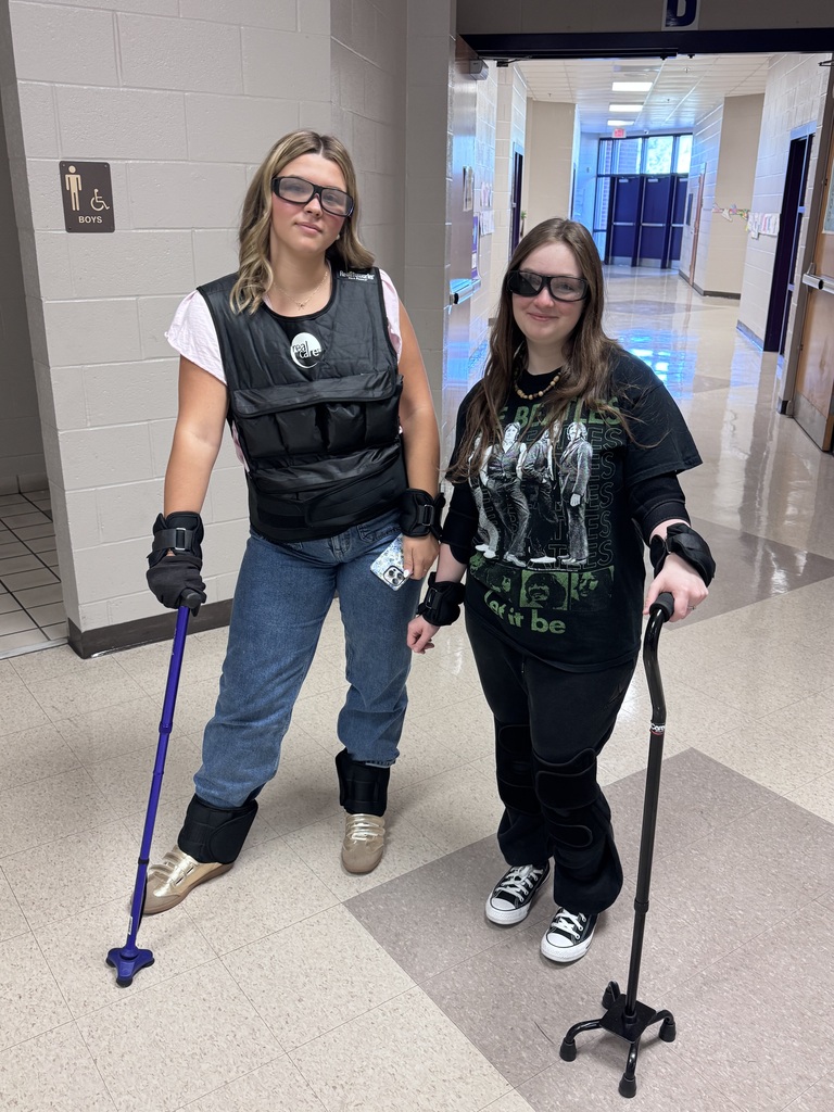 Two students wear geriatric impairment simulation gear with dark glasses and mobility canes in a school hallway.