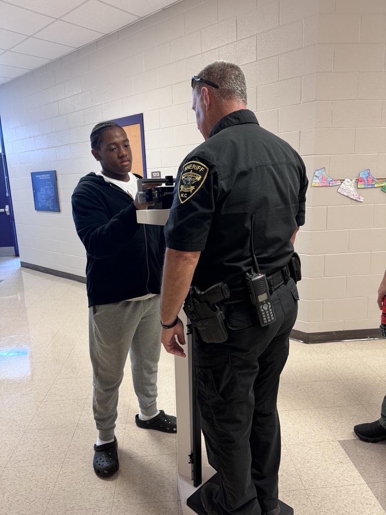 A uniformed sheriff's officer assists a student standing on a physician balance scale in a school hallway.