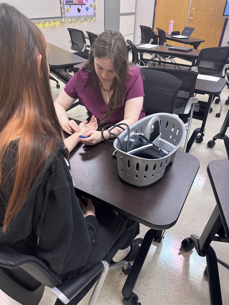 A student in scrubs checks another student's pulse at a desk with a blood pressure cuff nearby