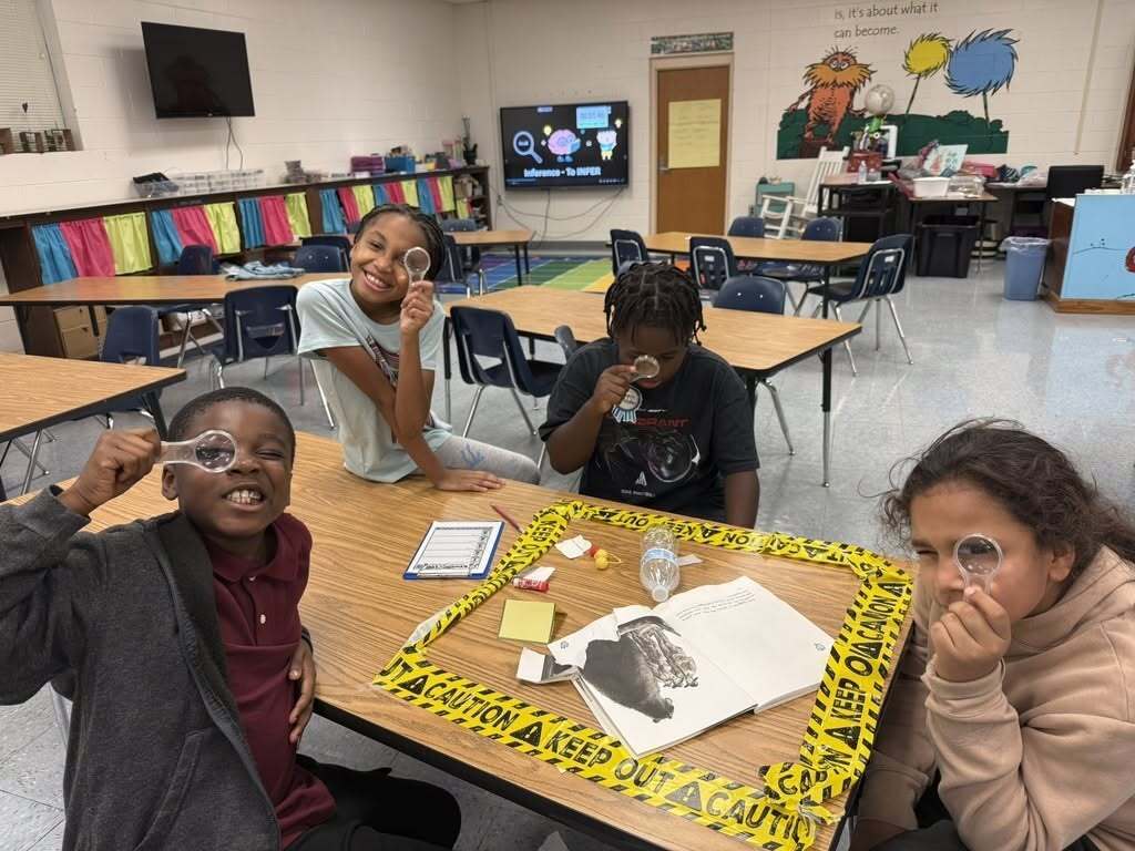 Four elementary students are using magnifying glasses while looking at library evidence at a table.