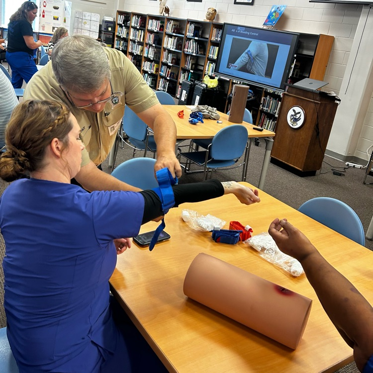 A lady in blue scrubs is checked by an officer to make sure she has correctly applied a tourniquet