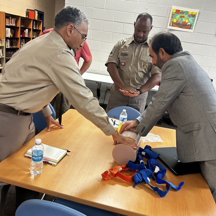 two males practice applying pressure to a mannequin while another gentleman stands by watching