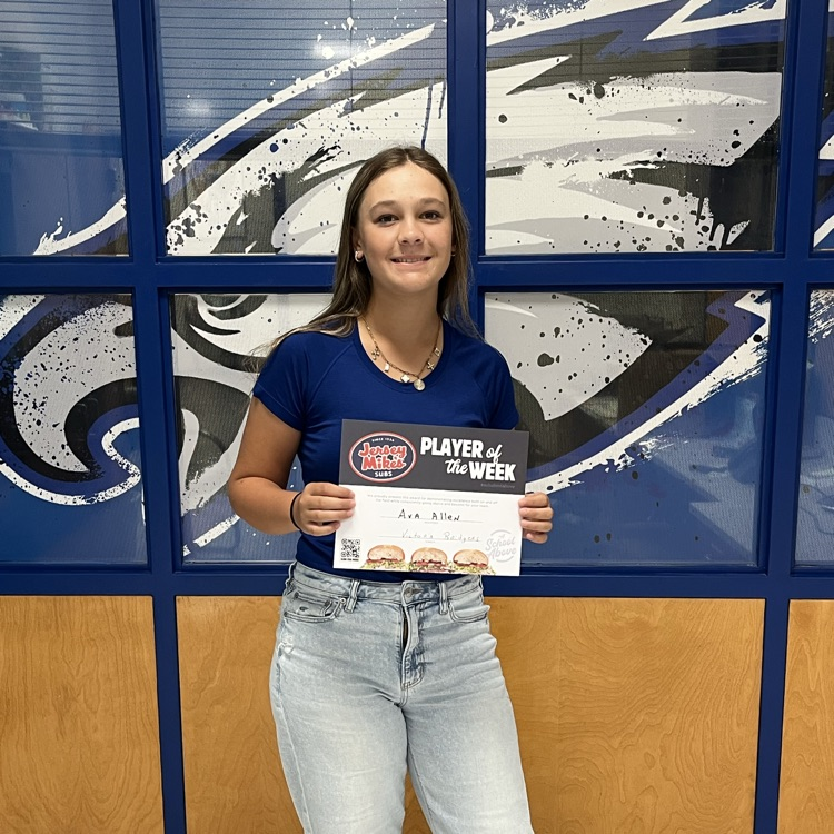 high school girl in light colored jeans, blue shirt, in front of windows with Eagle graphics, holding a Jersey Mikes certificate