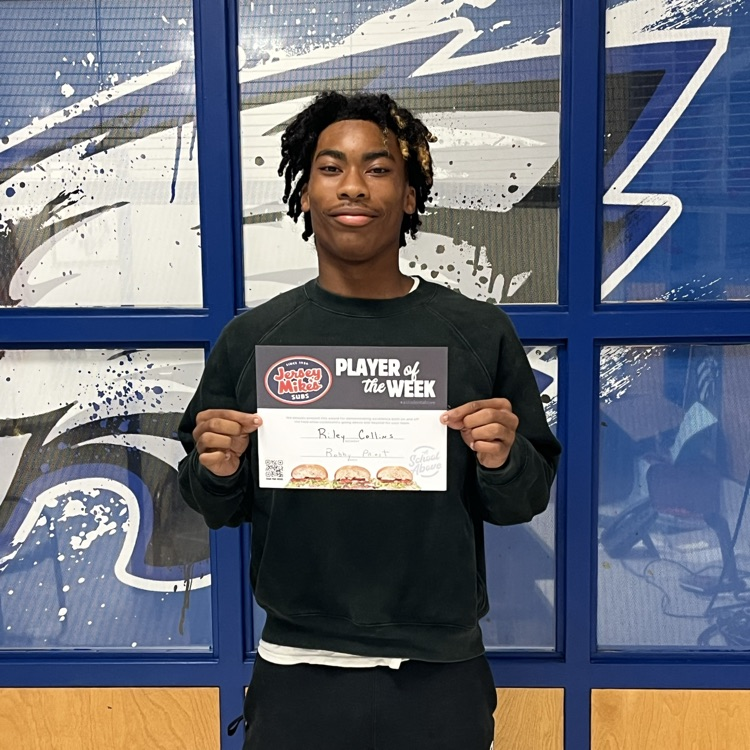 male high school student in black pants, black shirt, in front of windows with Eagle graphics, holding a Jersey Mikes certificate