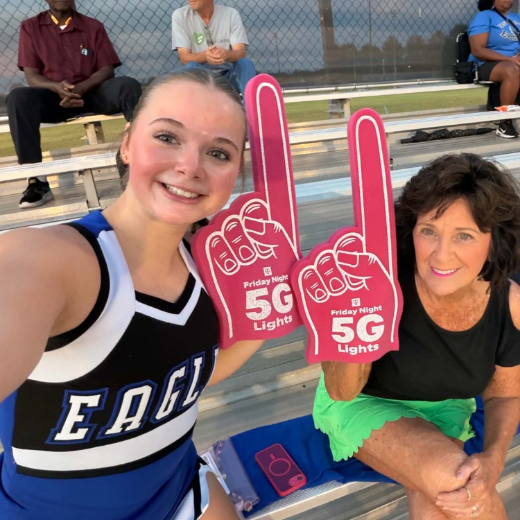 cheerleader in blue black and white uniform and lady in green shorts and black shirt posing with pink 5G foam fingers