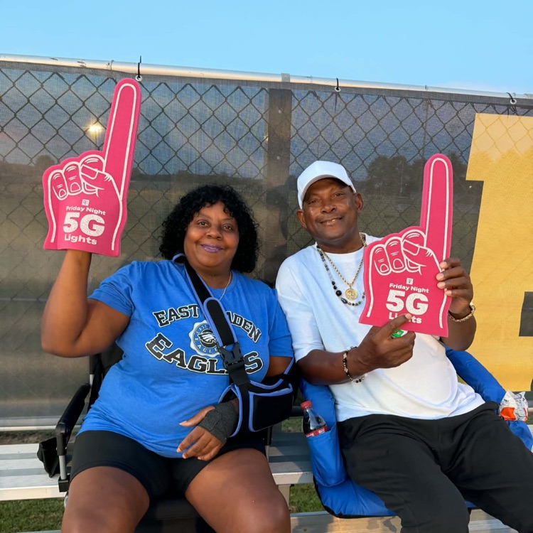 Woman in blue eagle shirt, black shorts and men and black pants white shirt posing with pink 5G, T-Mobile phone fingers that football game