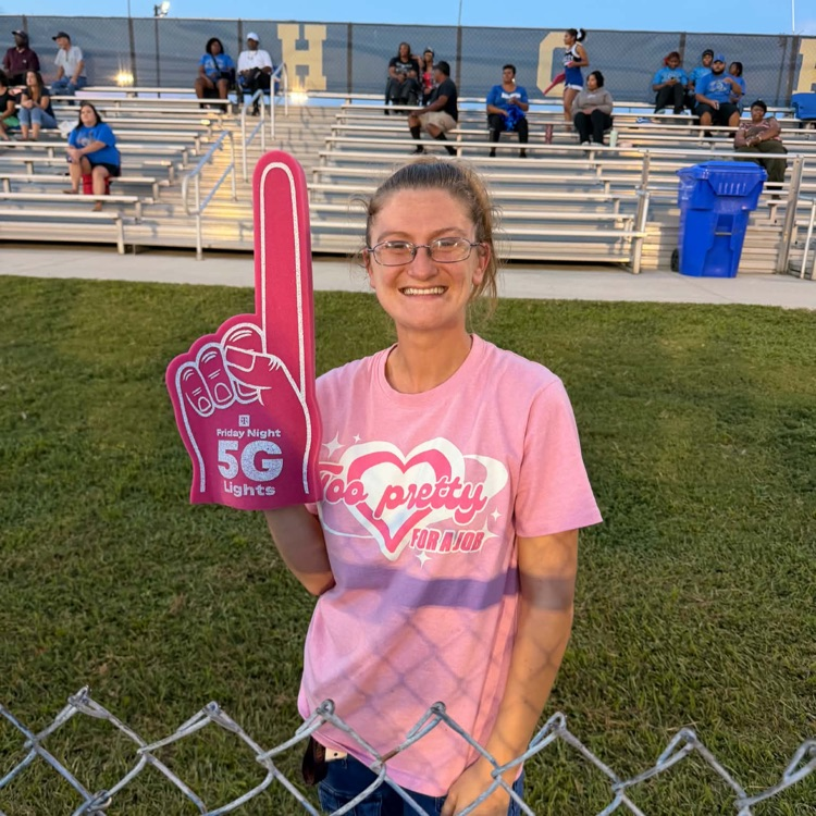 Lady in pink shirt poses on sidelines with pink T-Mobile from finger there are fans and bleachers behind her