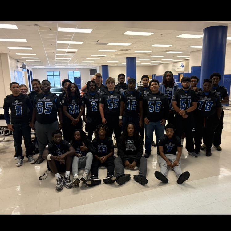 Group of football players in various bottoms with black and blue football jerseys, standing in Rose, and five sitting on the floor in the front in a cafeteria