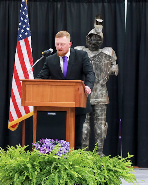 Coach Jackson speaking at the athletics banquet, dres in a dark suit and purple tie, standing at a podium with a knight and American flag in the background