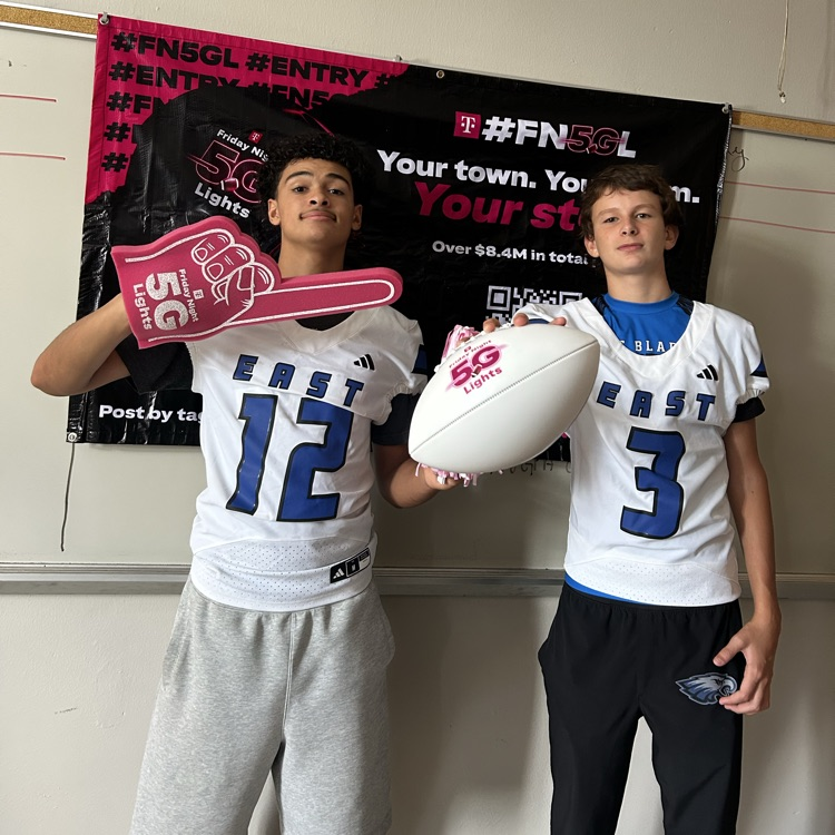 I football player in a white jersey and gray sweats holding a pink foam finger and a football player and a white jersey with black pants, holding a pink and white football in front of the T-Mobile Friday night lights banner!