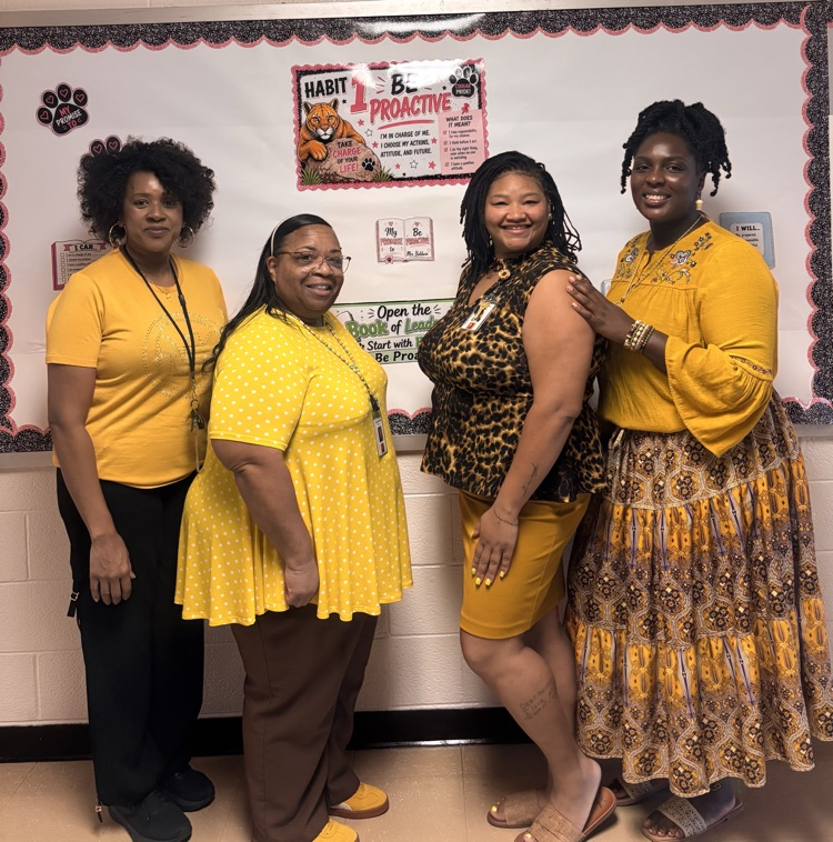 Four Teachers smiling wearing gold and yellow, standing in hall in front of bulletin board.