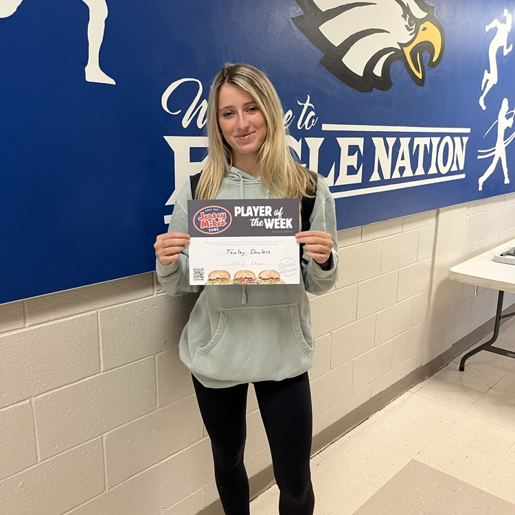 female with tan hoodie, black leggings in front of a white wall with a blue sign that says eagle nation with a eagle student is holding a Jersey Mike’s certificate