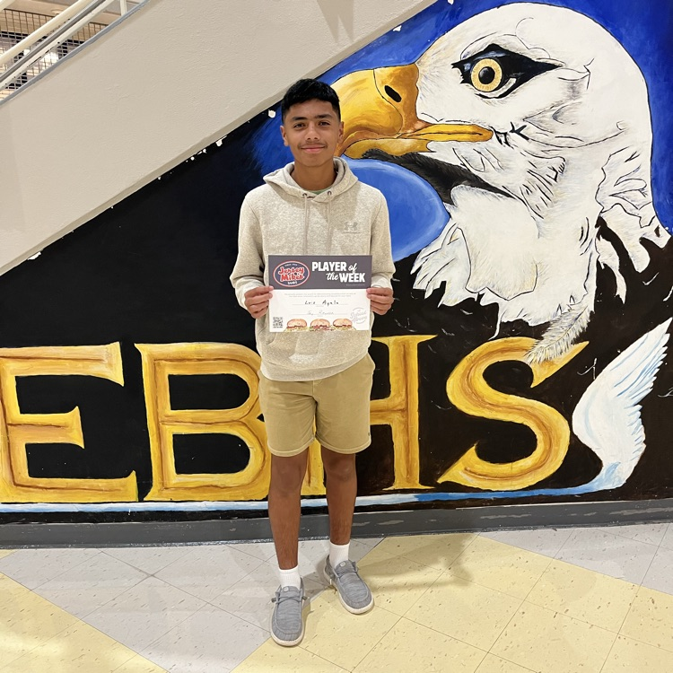 student with khaki shorts, white hoodie in front of a EBHS eagle background holding a Jersey Mike’s certificate