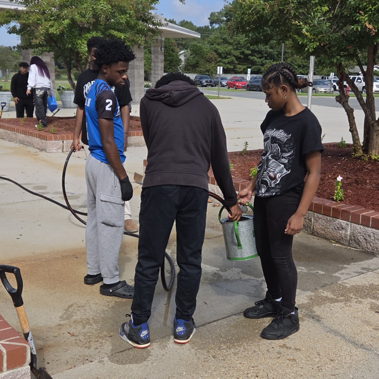 my student don’t hold a watering. Can while two other students help hold a hose and fill the can up. They are standing on it, and there are garden beds behind them.