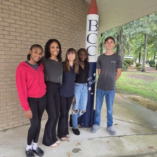 4 girls and 1 male standing on a sidewalk in front of a brick building and posing with a rocket looking statue that says "BCC"