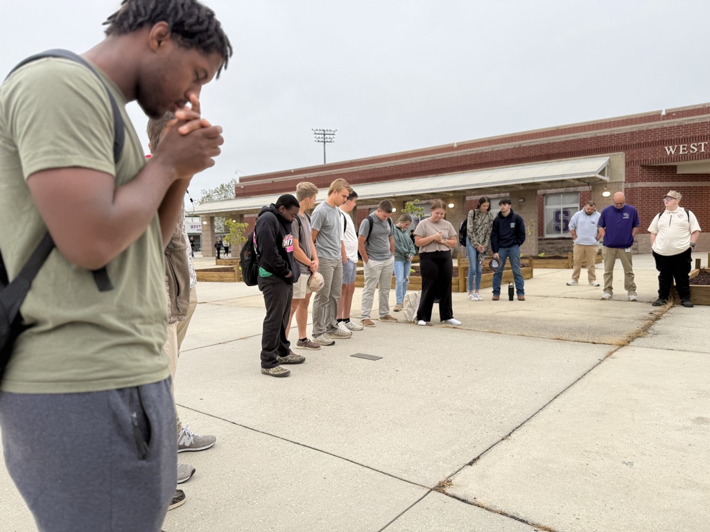 A student prays in the foreground outside West Bladen High School with a group of peers gathered in the background.