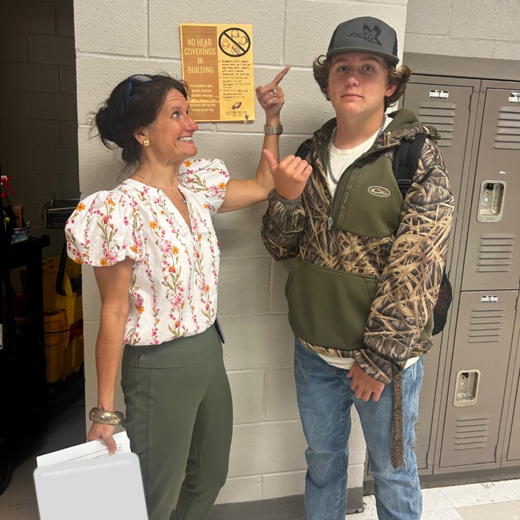 lady stands pointing to a hat on a student as he points to a no hat sign