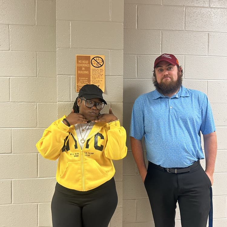 girl in yellow and man in blue wearing hats by an orange no hat sign 