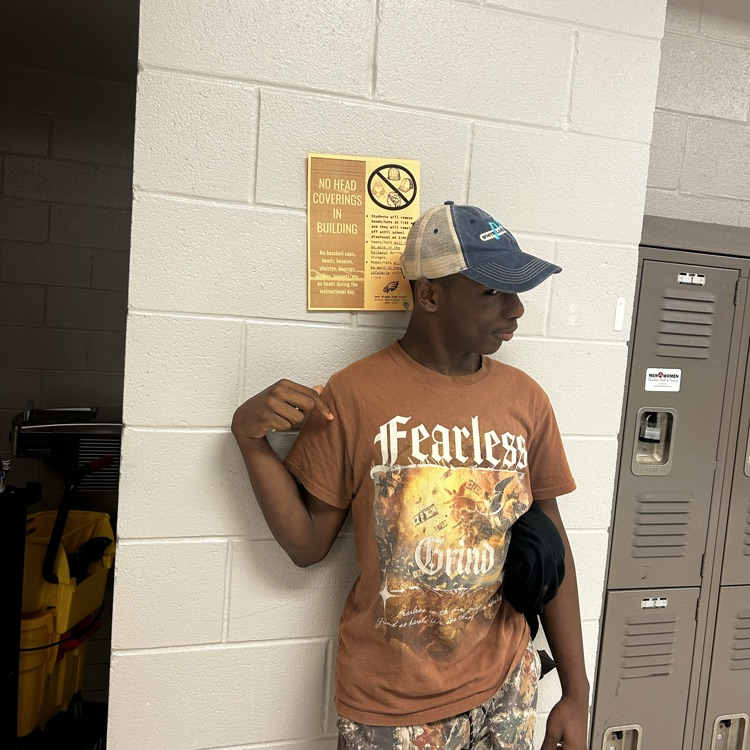 boy in camp pants and brown shirt wearing a hat by an orange no hat sign