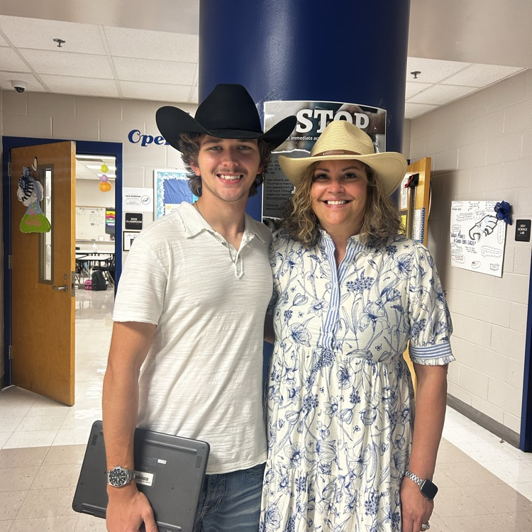 Lady and flowery dress with a boy and a white shirt, both wearing cowboy hats in front of a blue pole